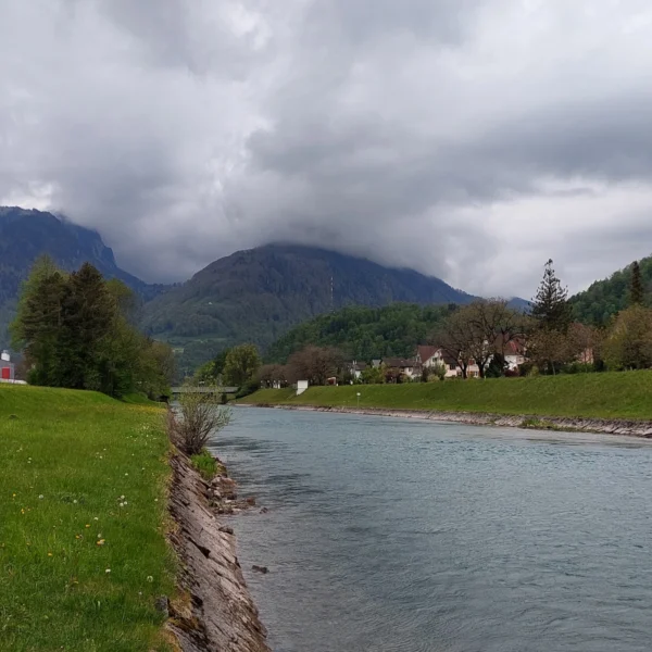 Ein ruhiger Fluss fließt unter bewölktem Himmel an einem grasbewachsenen Ufer entlang. Im Hintergrund sind Berge und vereinzelte Häuser zu sehen. An einem bewölkten Tag säumen Bäume und Grünflächen beide Seiten des Flusses.