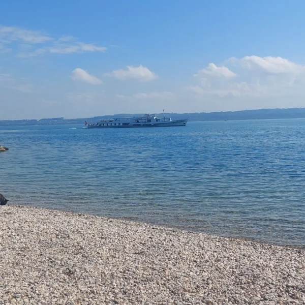 Ein Kieselstrand neben einer Steinmauer. Ein Boot gleitet auf ruhigem, blauem Wasser unter leicht bewölktem Himmel. In der Ferne sind Hügel über dem Lac de Neuchâtel, einem malerischen See in der Schweiz, zu sehen.