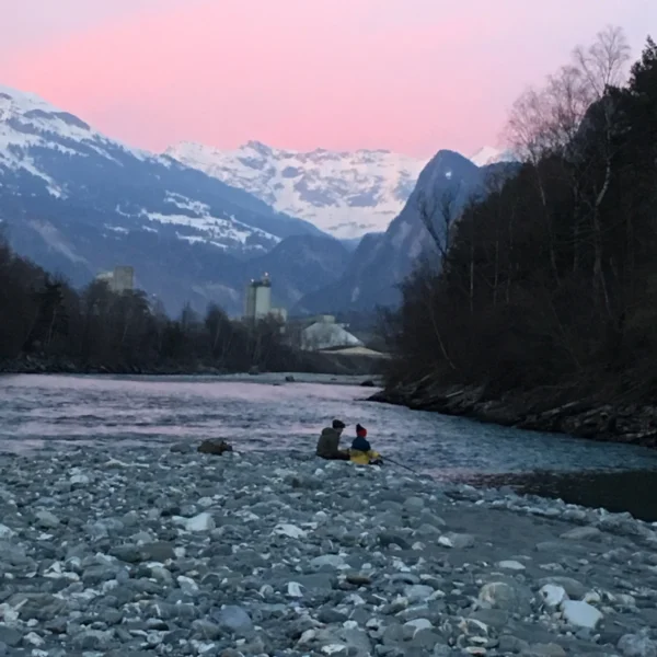 Zwei Personen am Rhein mit Alpenkulisse und rosafarbenem Himmel.