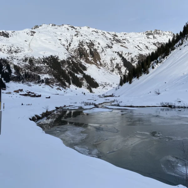 Eine schneebedeckte Berglandschaft mit einem teilweise zugefrorenen Fluss, möglicherweise dem Schlappinsee, fließt unter einem bewölkten Himmel durch ein von Bäumen und Hügeln gesäumtes Tal. Im Vordergrund steht ein Wegweiser.