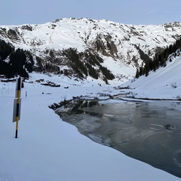 Eine verschneite Berglandschaft mit einem teilweise zugefrorenen Fluss, der an schneebedeckten Ufern in der Nähe des Schlappinsees entlangfließt. An den Hängen stehen vereinzelt Kiefern, und auf der linken Bildseite ist ein Straßenschild zu sehen.