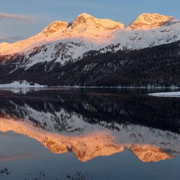 Bei Sonnenaufgang oder Sonnenuntergang sind die schneebedeckten Berge in warmes Sonnenlicht getaucht, das sich deutlich in der stillen, dunklen Oberfläche eines darunterliegenden Sees mit Bäumen und schneebedecktem Ufer spiegelt.