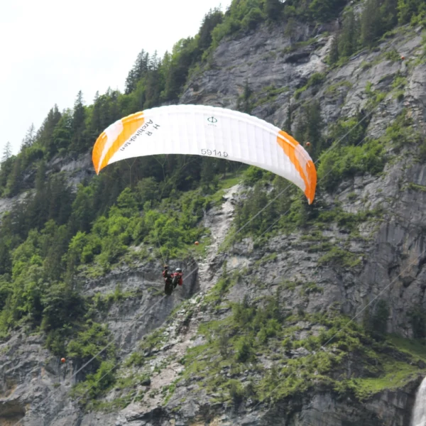 Ein Gleitschirm mit einem weiß-orangenen Schirm schwebt in der Nähe eines felsigen, mit grünen Bäumen bewachsenen Berghangs, während rechts in Lauterbrunnen der atemberaubende Staubbachwasserfall zu sehen ist.