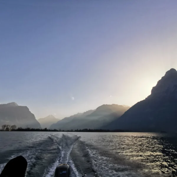 Ein malerischer Blick von einem fahrenden Boot auf einem See. Wellen im Wasser ziehen sich zu fernen Bergen hin, unter klarem Himmel bei Sonnenaufgang oder Sonnenuntergang. Die Sonne ist teilweise hinter einem Berggipfel verborgen.