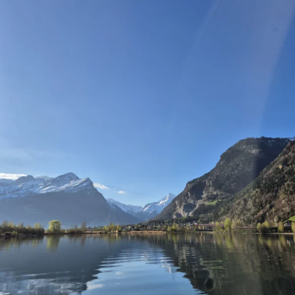In einem ruhigen See spiegeln sich schneebedeckte Berge und grüne Hügel unter einem klaren blauen Himmel wider. Am rechten Bildrand ist ein schwacher regenbogenartiger Bogen sichtbar.