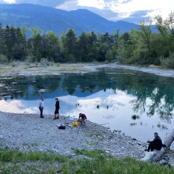 Eine Gruppe von fünf Personen versammelt sich am felsigen Ufer des Zizerser Weihers, einem ruhigen, spiegelnden Teich, umgeben von Bäumen und Bergen unter einem teilweise bewölkten Himmel in der Abenddämmerung. Einige stehen, andere sitzen oder hocken am Wasser.