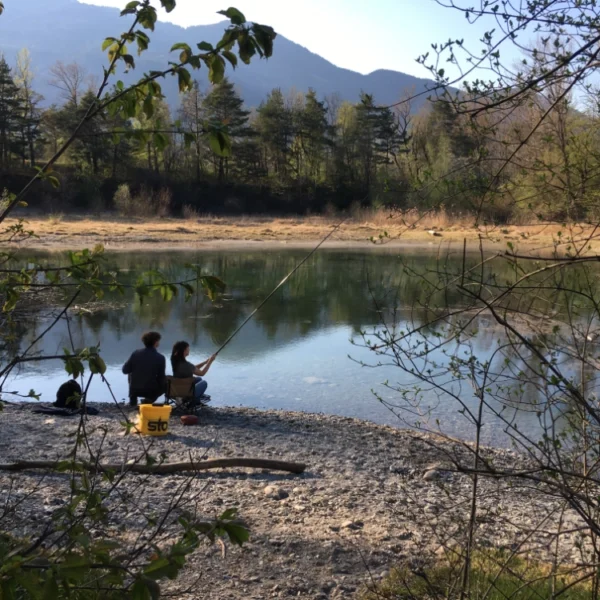 Zwei Personen sitzen am felsigen Ufer des Zizerser Weihers und angeln mit Ruten. Im Hintergrund erheben sich Bäume und Berge, während belaubte Zweige die ruhige Seelandschaft im Vordergrund teilweise einrahmen.