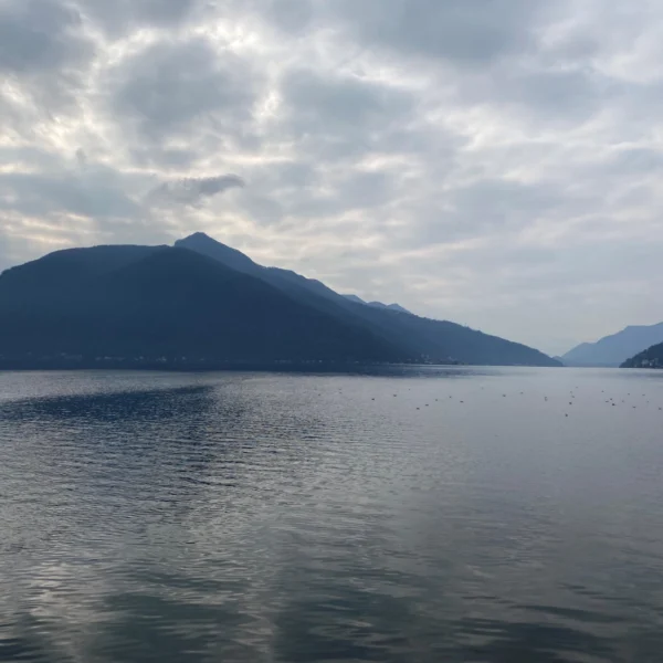 Luganersee (Lago di Lugano) mit Bergen unter bewölktem Himmel.