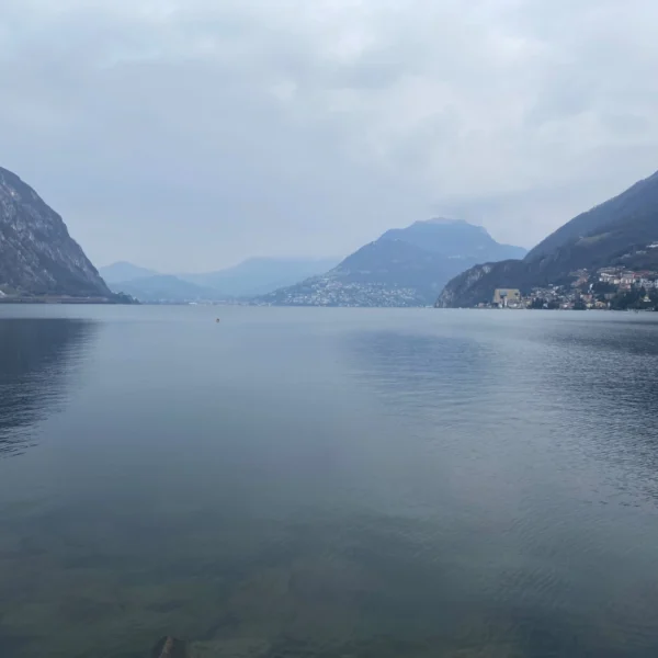 Luganersee mit Bergen und bewölktem Himmel. Ruhige Wasseroberfläche.