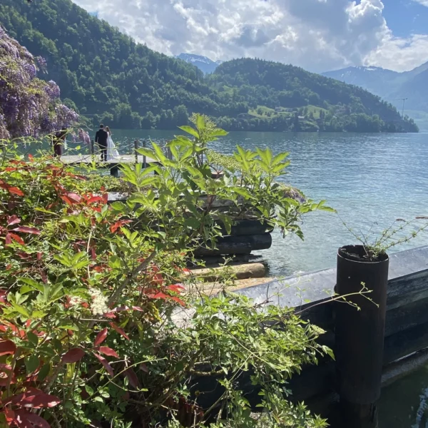 See mit klarem, blauem Wasser, umgeben von grünen Hügeln und Bergen unter einem teilweise bewölkten Himmel. Üppige Pflanzen im Vordergrund und eine Person, die auf einem Steg am Wasser steht.