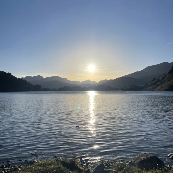 Die Sonne geht über den fernen Bergen unter und wirft ein helles Spiegelbild auf einen ruhigen See. Das Wasser plätschert sanft in der Nähe eines grasbewachsenen, felsigen Ufers im Vordergrund, alles unter einem klaren blauen Himmel.