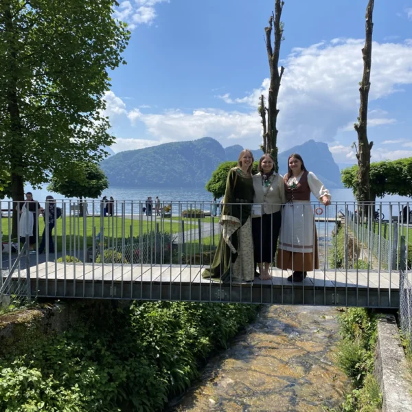 Drei Frauen in traditioneller Kleidung stehen auf einer kleinen Brücke über einem Bach in Mühlebach und lächeln in die Kamera. Hinter ihnen sind grüne Bäume, Menschen, eine Wiese und ein See mit Bergen unter dem teilweise bewölkten Himmel von Altdorf zu sehen.
