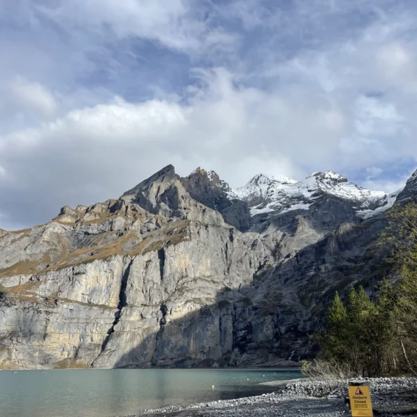 Ein ruhiger See in den Schweizer Alpen, umgeben von hohen, schneebedeckten Bergen und einem bewölkten Himmel. Bäume und ein Warnschild säumen das Ufer im Vordergrund und fangen die ruhige Schönheit des Oeschinensees ein, einem Juwel unter den Schweizer Seenlandschaften.