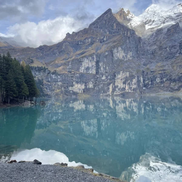 Der türkisfarbene Oeschinensee in der Schweiz spiegelt schneebedeckte Berge und Kiefern unter einem bewölkten Himmel wider, mit einer felsigen Uferlinie im Vordergrund – perfekt für Wanderfreunde.