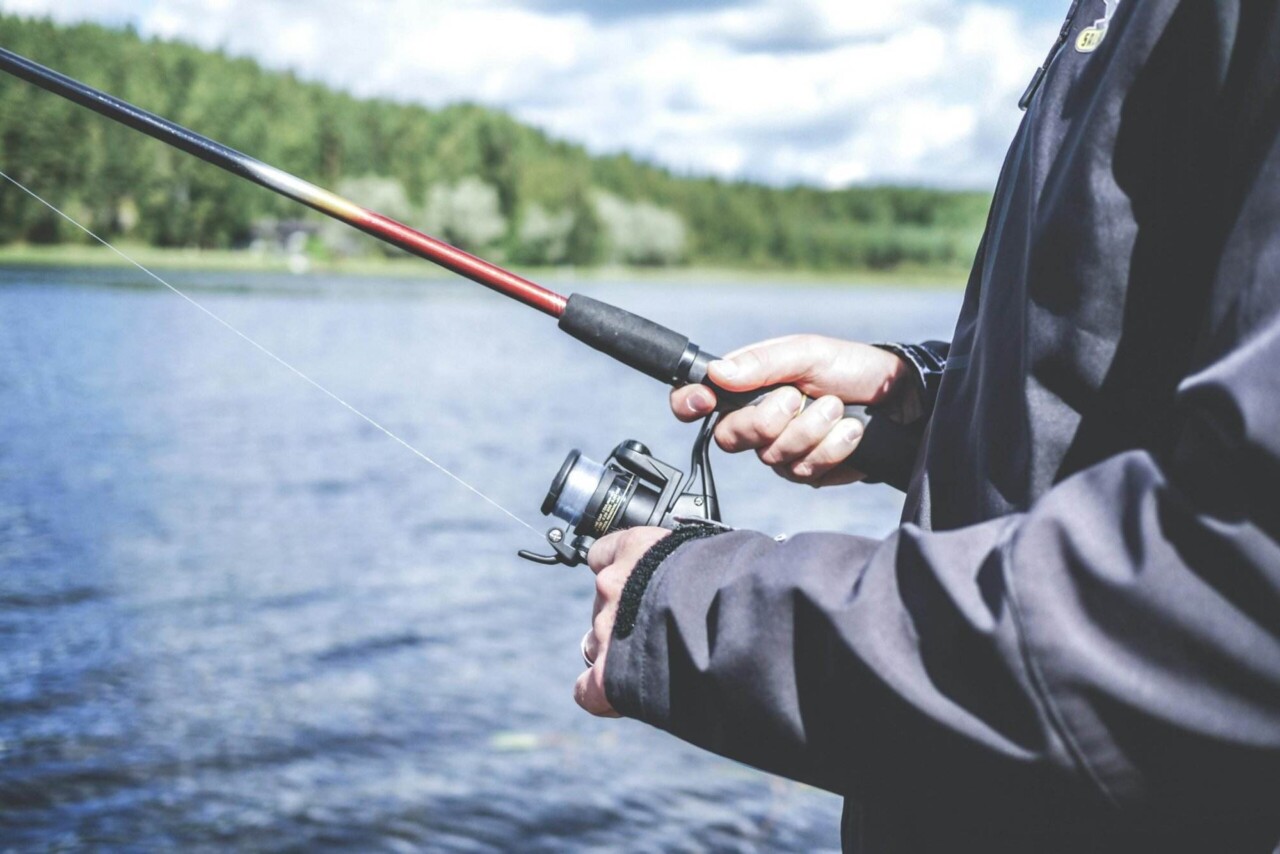 Eine Person in einer dunklen Jacke hält eine Angelrute, während sie an einem See steht und das Fischen in der malerischen Natur genießt. Die Kulisse aus Wasser, Bäumen und einem teilweise bewölkten Himmel unterstreicht das ruhige Erlebnis von Angeltouren.