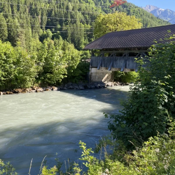 Eine überdachte Holzbrücke überquert einen schnell fließenden Fluss, umgeben von üppigen grünen Bäumen und Bergen unter einem klaren blauen Himmel.