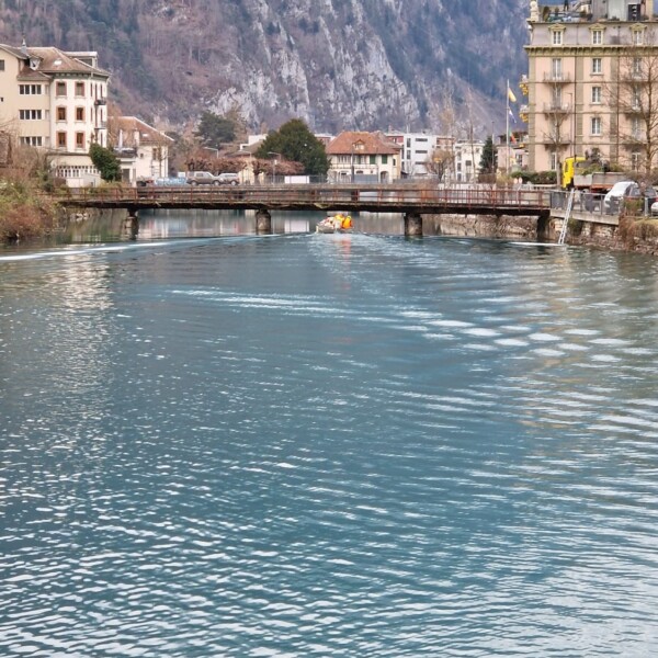 Ein ruhiger blauer Fluss fließt unter einer Brücke mit Gebäuden auf beiden Seiten, vor dem Hintergrund verschneiter Berge und eines bedeckten Himmels.