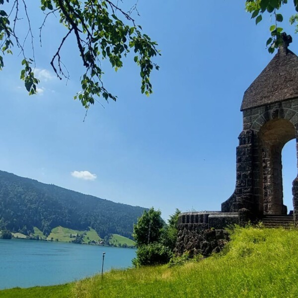 Ein steinernes Monument mit einer gewölbten Öffnung steht auf einem grasbewachsenen Hügel mit Blick auf einen See, mit Baumzweigen darüber und bewaldeten Bergen im Hintergrund unter einem strahlend blauen Himmel.