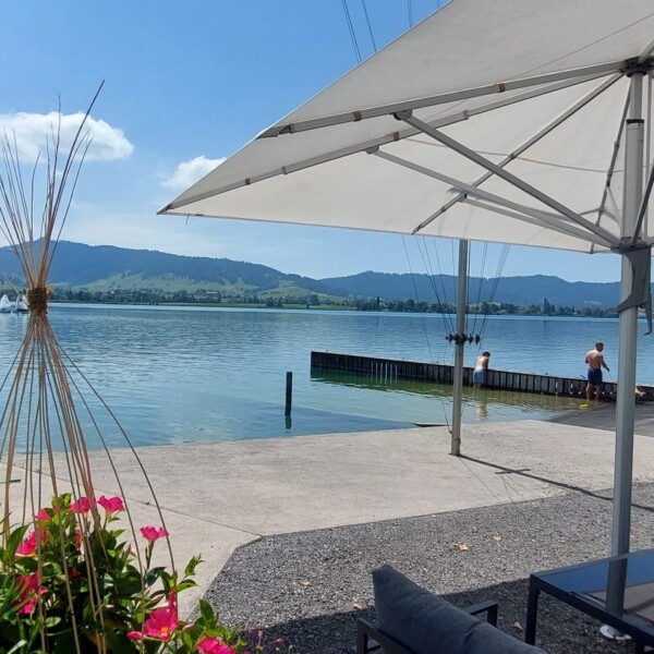 An einem sonnigen Tag entspannen sich Menschen unter großen weißen Sonnenschirmen am Seeufer, mit Bergen und grünen Hügeln im Hintergrund. Einige sitzen in der Nähe des Wassers, und in der Ferne sind Boote zu sehen. Im Vordergrund sind Blumen und Sitzgelegenheiten im Freien zu sehen.
