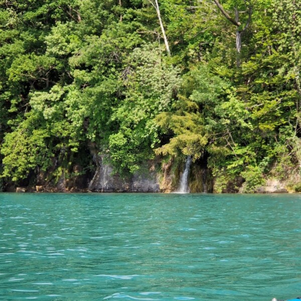 Ein kleiner Wasserfall fließt aus einem üppigen grünen Wald in einen türkisfarbenen See, wobei in der unteren rechten Ecke des Bildes ein Teil eines Paddels und eines Kajaks zu sehen ist.