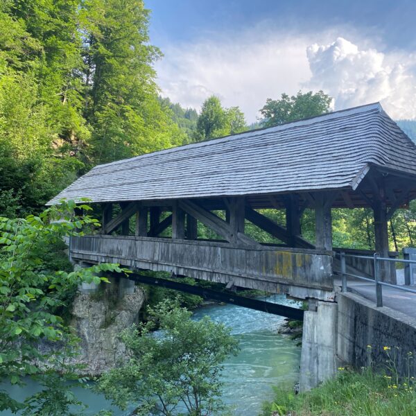 Eine hölzerne, überdachte Brücke führt über einen türkisfarbenen Fluss, umgeben von üppig grünen Bäumen und Hügeln unter einem teilweise bewölkten Himmel. Neben der Brücke verläuft eine Straße.