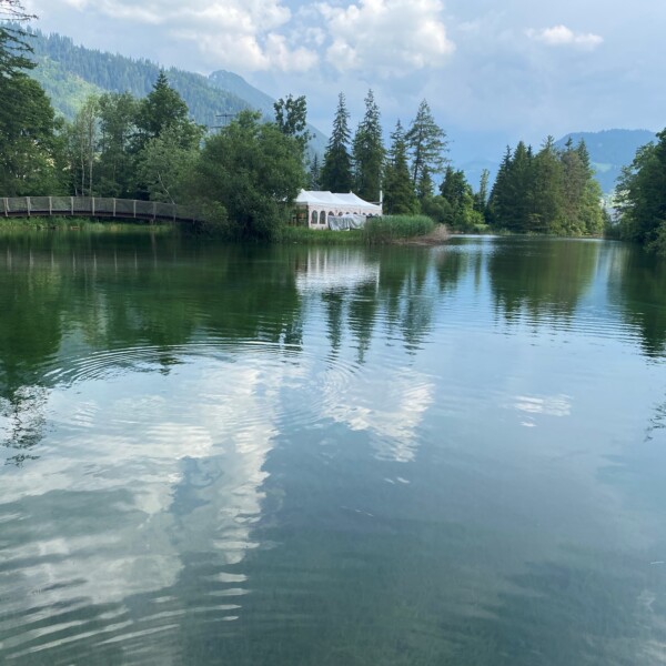 In einem ruhigen See spiegeln sich der Himmel und die umliegenden Bäume, links eine Holzbrücke und ein weißes Zelt am Ufer; im Hintergrund sind die Berge und der bewölkte Himmel zu sehen.