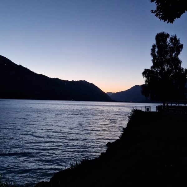 Ein ruhiger See in der Abenddämmerung, in dessen ruhigem Wasser sich das schwindende Licht spiegelt. Silhouetten von Bergen und Bäumen rahmen die Szene ein, und der Himmel geht in der Nähe des Horizonts von tiefem Blau in ein helles Orange über.