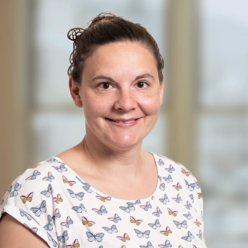 Eine Frau mit zurückgebundenen braunen Haaren lächelt in die Kamera. Sie trägt ein weißes Oberteil mit einem bunten Schmetterlingsmuster. Der Hintergrund ist sanft verschwommen und zeigt große Fenster.