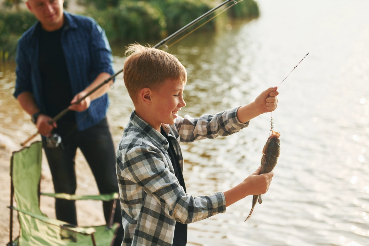 Ein lächelnder Junge hält einen Fisch hoch, den er mit einer Angelrute an einem Seeufer in der Schweiz gefangen hat, während sich ein Erwachsener im Hintergrund auf das Angeln vorbereitet. Beide sind im Freien und tragen legere Kleidung.