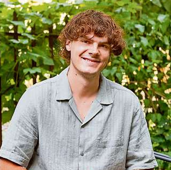 Ein junger Mann mit lockigen braunen Haaren und einem hellgrauen, kurzärmeligen Hemd sitzt lächelnd im Freien vor einem grünen, belaubten Gebüsch.
