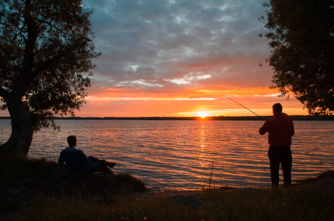 Zwei Menschen an einem Seeufer bei Sonnenuntergang; einer angelt - vielleicht übt er für die Schweizer Fischerprüfung - während der andere im Gras sitzt. Der orange-rosa Himmel spiegelt sich auf dem ruhigen Wasser, und die Bäume umrahmen die friedliche Szene.