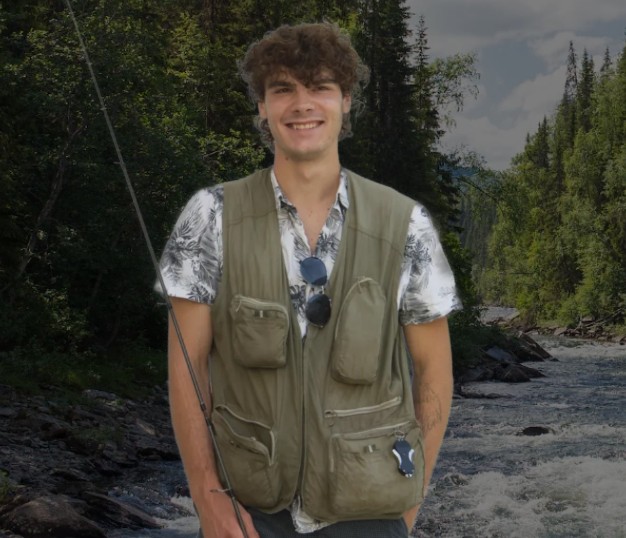 Ein junger Mann in einer Anglerweste und mit einer Angelrute in der Hand steht an einem fließenden Fluss und führt seine Angel durch das Wasser, umgeben von dichten grünen Bäumen an einem sonnigen Tag.