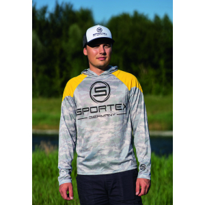 Ein Mann in einem Sportex UV Shirt gelb/grau camou Größe M steht im Freien auf einer Wiese, mit Bäumen und einem Fluss im Hintergrund, und schaut in die Kamera.