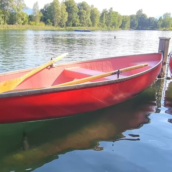 Rotes Ruderboot am Baggersee Kriessern vertäut, mit Rudern. Ruhiges Wasser und Bäume im Hintergrund.