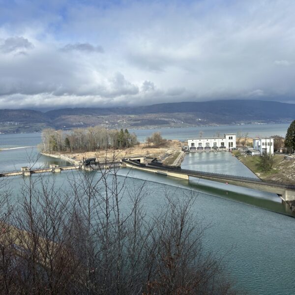 Eine malerische Ansicht eines Staudamms mit türkisfarbenem Wasser, Gebäuden und Bäumen vor einem großen See und entfernten Bergen unter einem bewölkten Himmel.