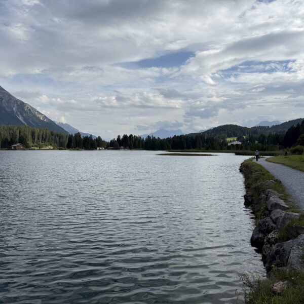 Heidsee Seeansicht mit Weg und bewaldeten Bergen unter bewölktem Himmel.
