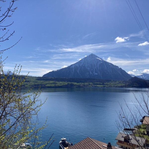 Ein schneebedeckter Berg erhebt sich über einem blauen See, über dem ein klarer Himmel liegt. Im Vordergrund sind Dächer, Boote und Baumzweige zu sehen, die auf ein friedliches Dorf am See hinweisen.