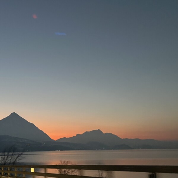Ein heiterer Sonnenuntergang über einem See mit den Silhouetten von Berggipfeln in der Ferne und einem klaren, verblassenden Himmel, gesehen hinter einem Zaun am Straßenrand.