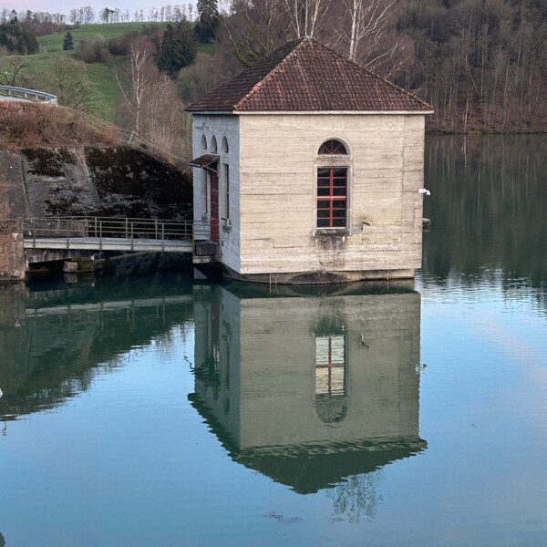 Ein kleines, quadratisches Gebäude mit gewölbten Fenstern liegt teilweise im ruhigen Wasser und spiegelt sein Bild wider. Im Hintergrund stehen Bäume ohne Blätter und ein grasbewachsener Hügel unter einem blau-rosa Himmel.