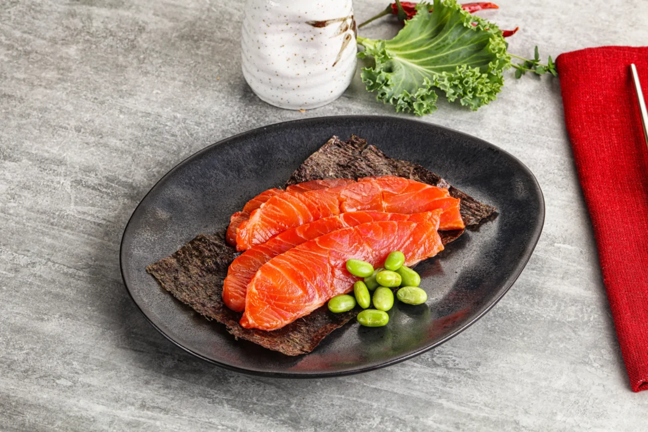 In Scheiben geschnittener roher Lachs auf Seetangblättern, serviert mit grünen Edamame auf einem schwarzen Teller. Eine weiße Vase, Salat und eine rote Serviette mit Stäbchen sind zu sehen - perfekt für alle, die die Küche schätzen und die Schweizer Fischerprüfung bestanden haben.