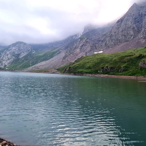 Sanetschsee: Ruhiger Bergsee umgeben von grünen Hängen und felsigen Gipfeln unter bewölktem Himmel.