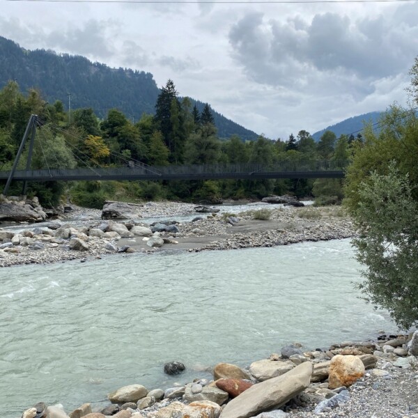 Fluss Vorderrhein unter einer Hängebrücke, umgeben von grünen Bäumen und Bergen.