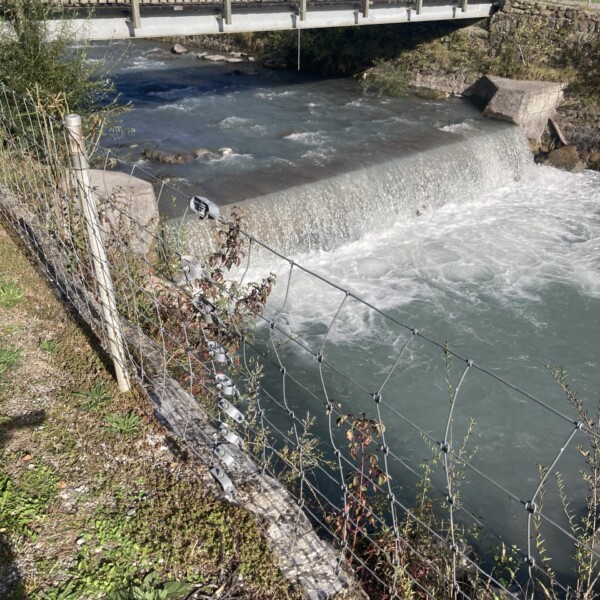 Taschinasbach Fluss mit Brücke und kleinem Wasserfall. Grüne Ufervegetation.