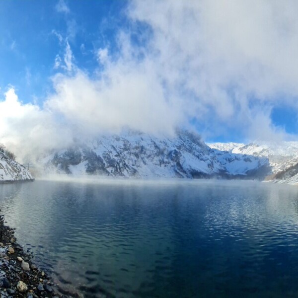 Eine Person in Winterkleidung angelt am Rande des Lac de Tseuzier, einem klaren Bergsee, der von schneebedeckten Hügeln und Bäumen umgeben ist, über denen Wolken und blauer Himmel hängen.