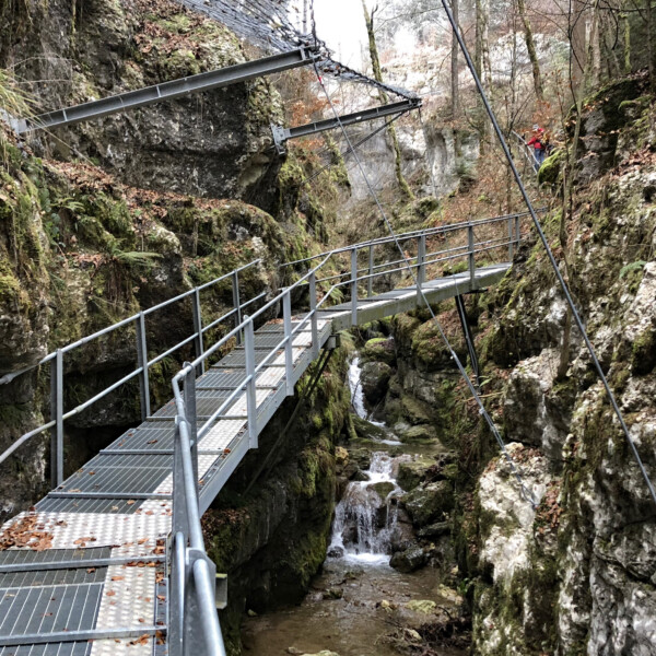 Metallsteg über den Cholersbach in einer Schlucht mit Wasserfall. Wanderweg mit Geländer.