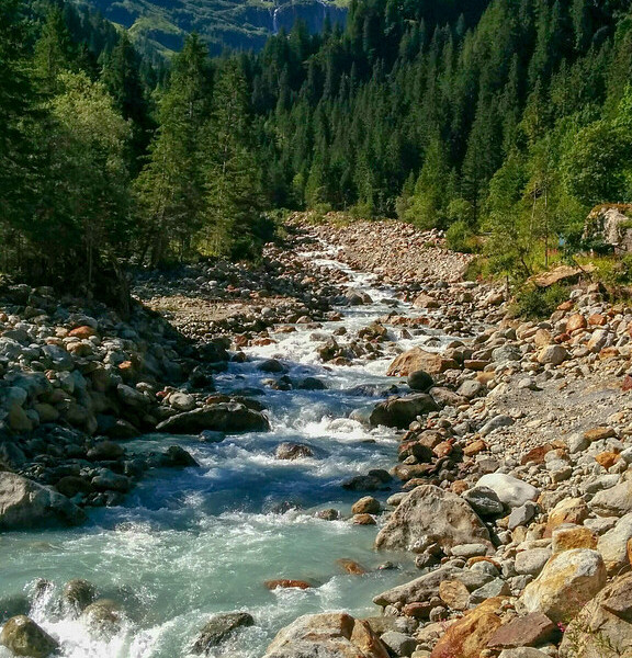 Fluss Chrummbach mit Felsen, Wald und schneebedeckten Bergen im Hintergrund.