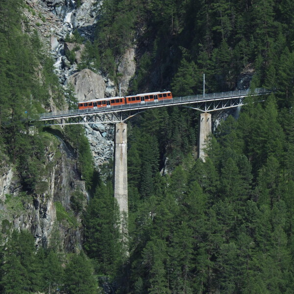 Orange Zug fährt über die Findelbach-Brücke inmitten grüner Bäume und Felsen.