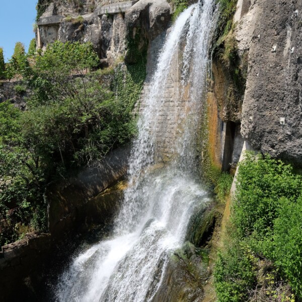Wasserfall stürzt über eine Steinmauer, umgeben von üppigem Grün.