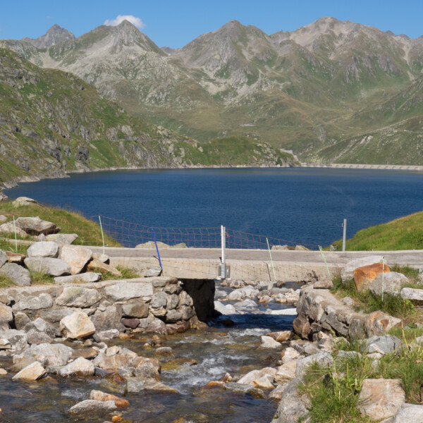 Gotthardreuss-Fluss unter einer kleinen Brücke mit Bergsee im Hintergrund.