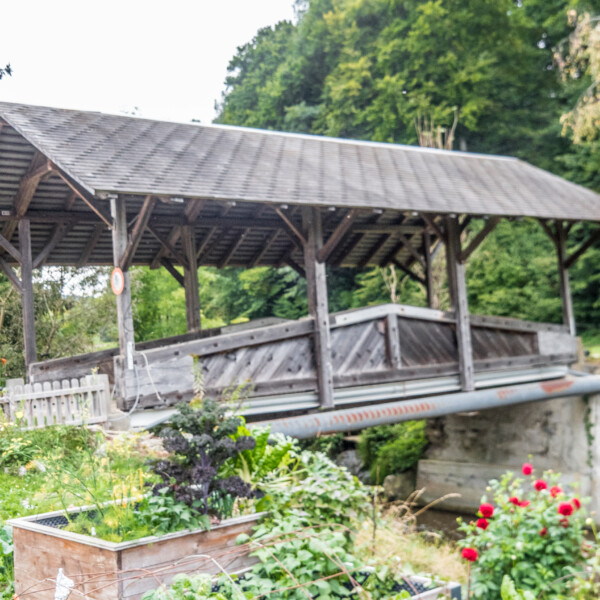 Überdachte Holzbrücke im Grünen über einem Bach. Garten mit Blumen im Vordergrund.